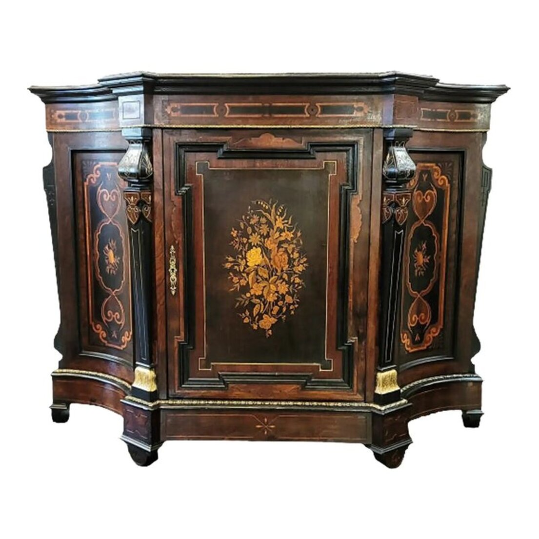 Victorian credenza and music cabinet has a shaped top with a molded edge enhanced with gilt bronze trim.  The frieze is decorated with marquetry strapwork. The bottom edge is enhanced with gilt bronze trim.  The case is made of maple wood with inlaid rosewood veneers and an ebony finish simulating Asian lacquer.  Two decorative supports are on the front of the credenza at either side of the single door cabinet at center. The supports are incised gilded and ebonized wood.  The central cabinet door is decorated with an intricate molded frame into which is set a rich marquetry panel of different exotic woods forming a bouquet of flowers. The interior of the door is paneled with birds-eye maple wood.  The inset hinges and lock are made of solid brass. The keyhole escutcheon on the door is in an openwork arabesque form.  The interior of the cabinet is paneled with maple wood and is fitted with a substantial solid maple wood removable shelf.  The concave side panels also feature rich marquetry motifs of musical instruments framed by anthemia.  Two more decorative supports are at each side back. They are incised and ebonized wood.  The base is shaped to reflect the top, has a molded top edge enhanced with gilt bronze trim, and an incised apron.  The four demi-lune feet are placed under molded top edges and feature incised decoration.  The back is framed panel constructed with an ebonized finish. Available from India Street Antiques, San Diego.