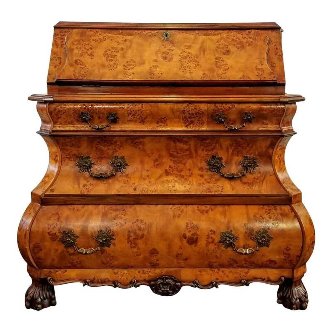 Dutch burr figure walnut wood bombe bureau in the Queen Anne / William and Mary style of the early 18th century.  A cyma curve slant drop front secretary desk is below the shaped top and opens via chased brass hinges to reveal serpentine pigeon holes and two small storage drawers with turned brass knobs. The small interior drawer construction features a nod to the late-17th and early-18th century practice of placing the drawer bottom wood grain front to back. The desk does lock and comes with a key. The key escutcheon features a fan and scroll motif executed in repousse brass.  Below that is a shaped top over cyma curve edged bombe three-drawer chest.  The three shaped drawers open by tugging on the cast brass rocaille bail pulls featuring Flemish scrolls, fans, and fuchsia flower motifs. The bail pull brass scutcheons feature a stylized chrysanthemum motif.  The carved apron at bottom features Flemish scrolls meeting in the center at a foliate cartouche.  The four feet are carved in a mid-to-late Queen Anne period hairy lion paw and ball style.  The back is an oak wood panel.  The desk was made post-World War II, in the 1950s and is a recreation of early-18th century styles popular during the William and Mary, Queen Anne, and Early Georgian periods. The bureau is very well constructed.  SIZE:  44