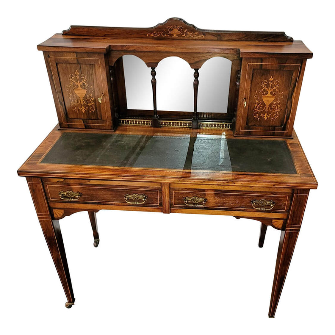 Late nineteenth century English rosewood writing table enriched with inlaid work by Maple & Company.  The curved board across the top is inlaid at the center with a satinwood urn and scrolled acanthus leaves.  The geometric shaped rosewood top rests upon two cupboards and an arcaded recess with a beveled mirror back, turned supports, and brass gallery.  The cabinet doors are inlaid with satinwood urns and acanthus arabesques. The doors lock and come with keys. The left cabinet interior features two standard shelves and the right cabinet interior features three vertical pigeon holes for correspondence.  The writing surface is inset with black leather embossed on the edges with a Greek key motif.  Below the writing surface are two frieze drawers constructed with fine hand-cut dovetailing and hand-planed bottoms. The drawer pulls are a bail type on geometric Aesthetic Movement pierced and chased brass scutcheons.  The square tapered legs end in brass casters.  Inlaid satinwood banding accents the cabinet doors, the writing surface, the drawer fronts, the legs, and the desk edges.  This model of writing table was sold by Maple & Company, England and was featured in their catalogue from 1885 to 1895.