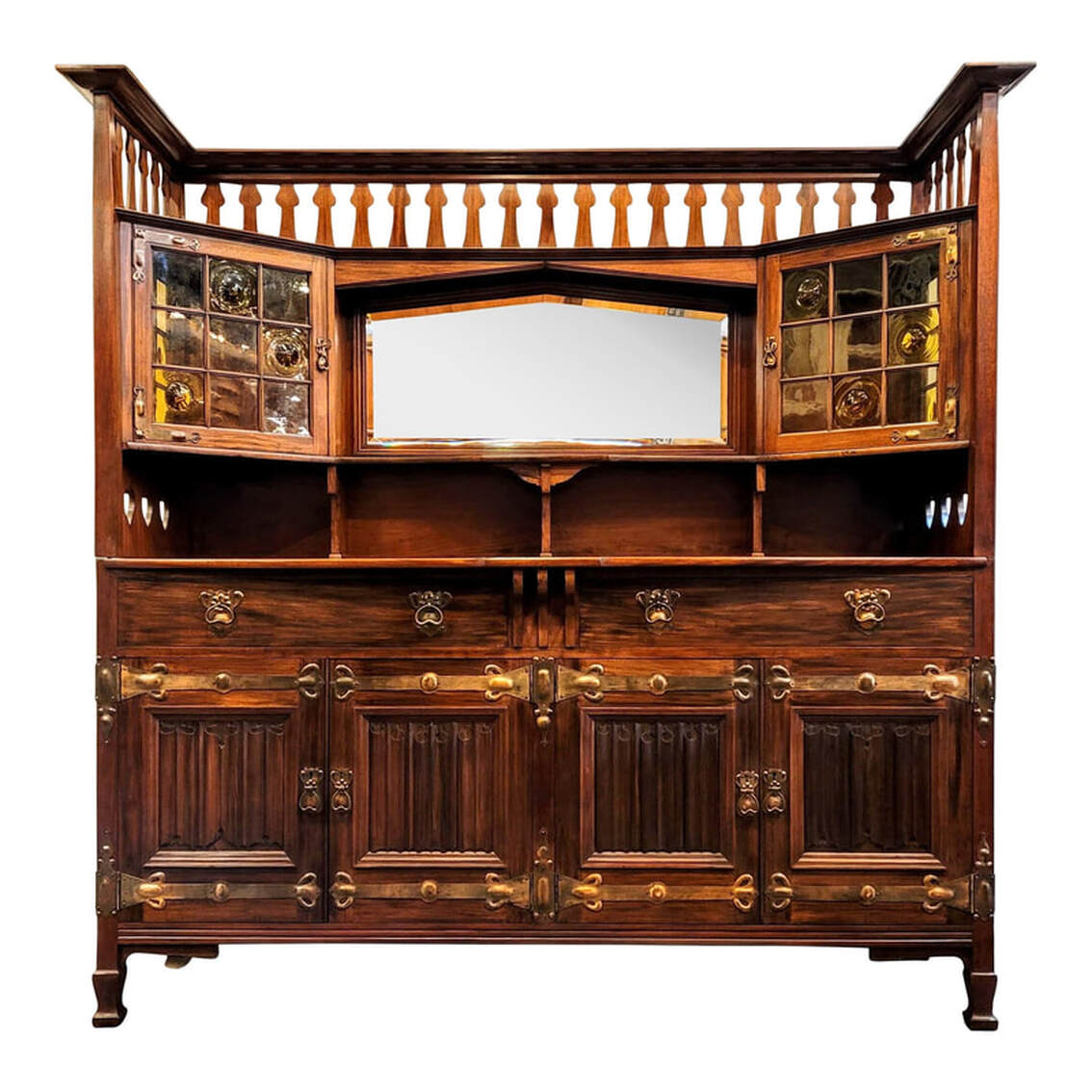 Shapland & Petter main sideboard 2130 in fumed West Indies mahogany consists of a buffet carcase with cellarette and a mirrored over-mantle with glazed cupboards.  The design of this sideboard dates from about 1894 through 1903. During this period, William Cowie was head of the design team which included James Henry Rudd and Henry Percy Shapland, grandson of the company founder.  The over-mantle consists of a molded flat top shelf surround, double grooved for platter and plate display. Below this is a pierced gallery of peacock fretwork. The central shaped beveled mirror is inset into a pointed arch frame - a distinct Shapland & Petter treatment used virtually exclusively as a regular feature - surrounded by molding. Under the mirror is a shaped shelf. Below the shelf are three shaped support brackets.  On each side is a canted cupboard with glazed doors and side panels. The glass panes are of a light amber-yellow tone. The plain panes are seedy glass and the remaining panes are blown crown antique bottle glass, or bullseye glass. The doors are decorated and attached with right angle corner pierced and hand-beaten repousse antiqued copper hinges. The drop pulls and pierced excutcheons are antiqued copper. The doors do lock and keys are provided. The inset brass lock on the left door is stamped 