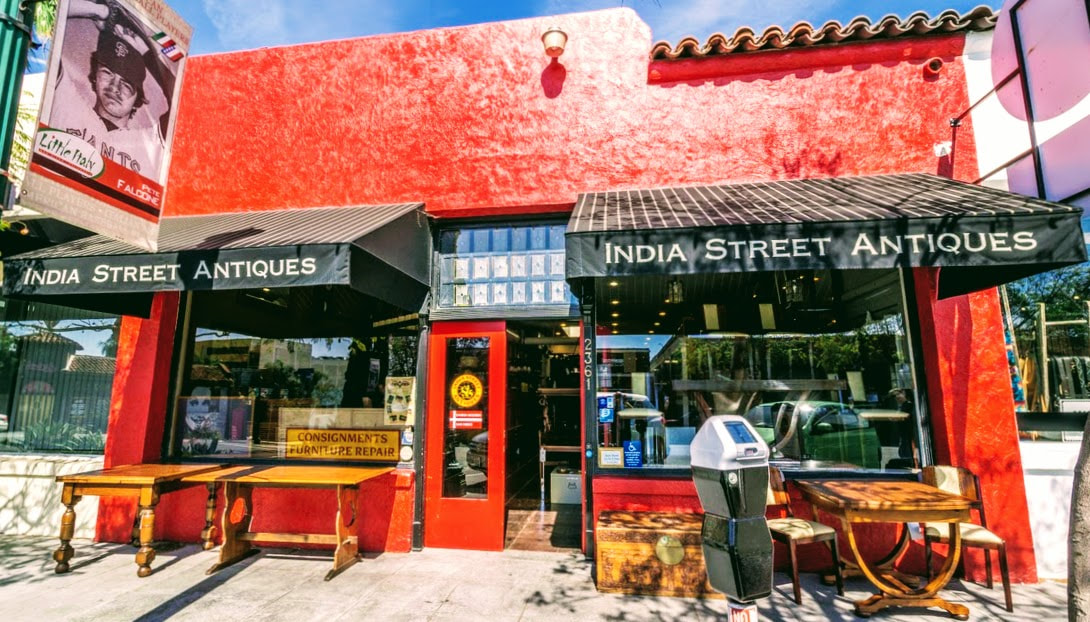 Photo by Noah Feldman of the India Street Antiques building at 2361 India Street, San Diego, 92101. The building is one story. painted red, with white-stripped black awnings over two large picture windows. Image is clickable to access the 710 San Diego Vacation Rentals article about India Street Antiques.