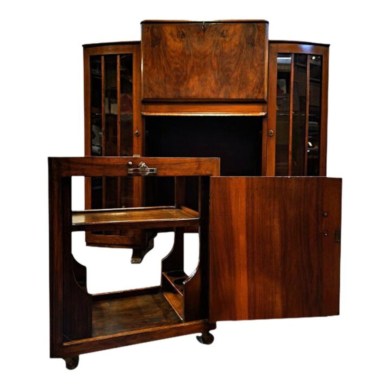 1920s drop-front butler's desk with side-by-side glazed cabinets and removeable bar cart was manufactured in England from burl figured walnut wood (Juglans Regia).  The flat top is leveled and has softly molded edges. The cabinets are topped with curved rear rails.  The drop-front desk is at center, top, and is fitted with pigeonholes separated by curvy dividers. There is storage space above and below the pigeonholes. The space at bottom may store a laptop and keyboard. The work surface on the back of the fall front door is inset with textured green leather. The desk does lock, and a key is provided. The desk sits high, as does a classic butler's desk, so it may be utilized as a standing desk or with a high stool.  Below the desk is a cabinet door that gives access to the installed bar cart; however, with a twist of the key and a press of the button, the bar cart pulls out completely and may be used as a separate piece of furniture, leaving space under the desk to store your work stoolor chair.  The walnut bar cart has a clear glass top above two wooden shelves. On the lower shelf are six holes for bottle storage, three on each side. The bar cart is on casters and rolls easily. The trolley may also be used as a coffee, tea, and cocoa station or as a space for your printer and other office equipment.  Two glazed display cabinets are fitted with two shaped glass shelves each. The cabinets lock and keys are included. These would house drinking glasses and decanters very well.  The keyhole escutcheons on the desk and the cabinet doors are shaped and marbled thermoplastic, similar to Catalin and Bakelite, in caramel, amber, and root beer tones.  The keyhole escutcheon on the bar cart is a scalloped roundel in brass.