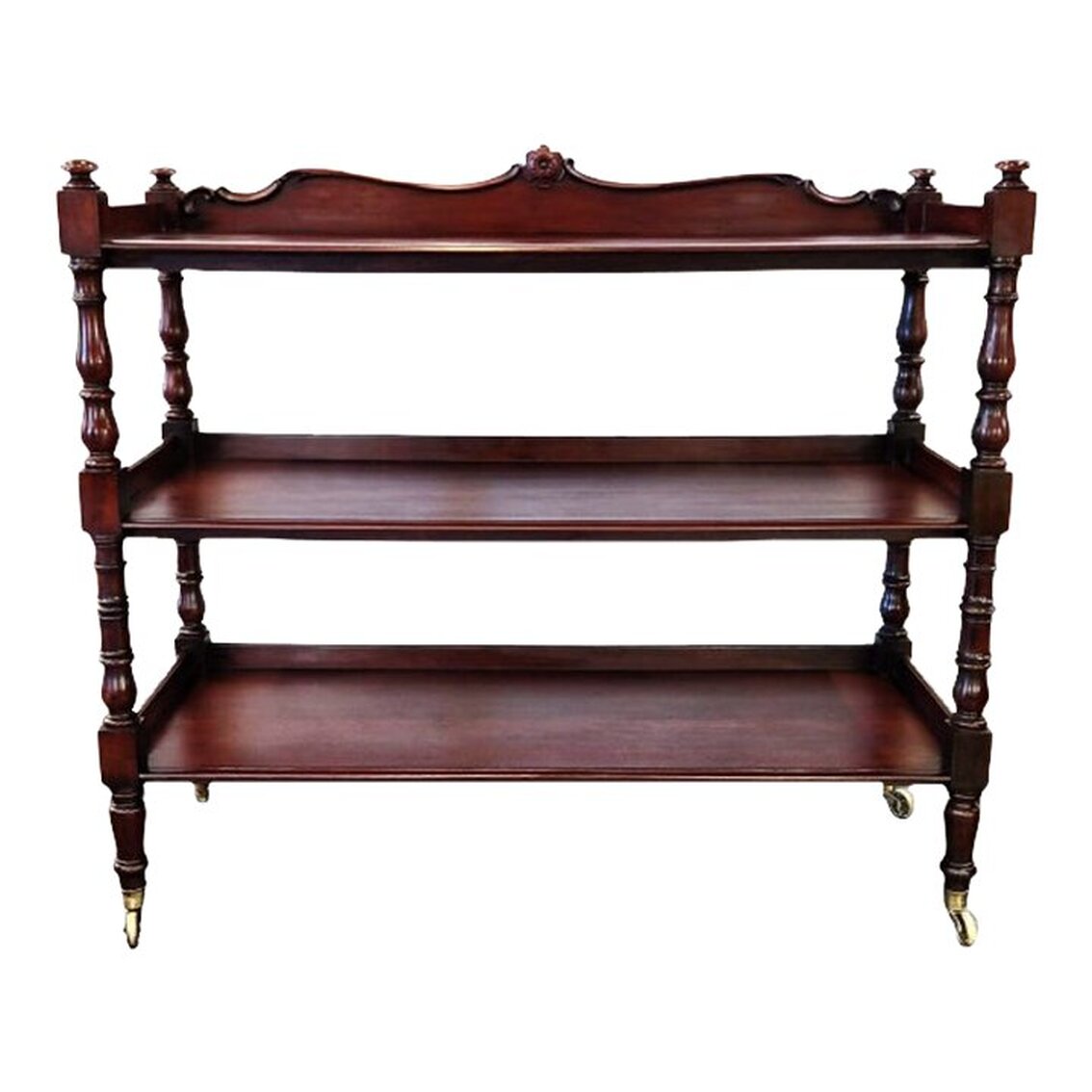 Antique circa 1820s-1830s food server is constructed of Caribbean mahogany wood and has three lipped shelves, four turned legs topped with knob finials ending in brass sabot casters, and a curved back rail with a central carved flower roundel.