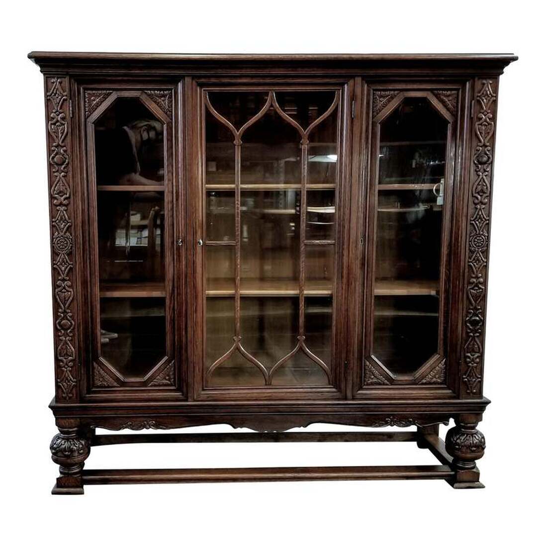 Glazed bookcase of carved oak wood in the Jacobean style. Three locking glass doors open to reveal wooden book shelves. Keys are provided for the locks. The carcass sits atop a base with low stretchers and is supported at the front corners by bulbous legs with acanthus leaf carvings. The stile strapping carvings combine bow & shield and floral & leaf designs.