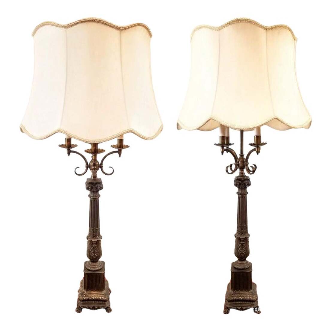 Pair tall metal table lamps in the French Empire candelabra style date to 1920-1926, a period of revival in this style of lighting.  The patinaed brass candelabra form features Neoclassical elements such as scalloped bobeche supported by fluted scrolls collared by bosses; acanthus fluted Ionic columns supported by laurel wreath surmounted plinths; and lion paw and claw feet.  Each lamp has a scalloped and trimmed hand constructed creamy white silk shade. The lighting is activated by three-way black rotary knob switches. The first rotation turns on just the candle flame bulbs; the second rotation turns on just the central light bulbs; the third rotations turns on all the lights; and the fourth rotation turns the lamps off. SIZE:  42.5