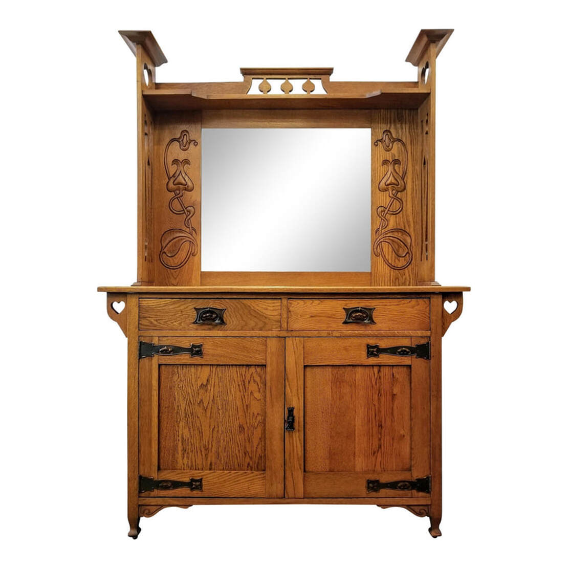 Antique main sideboard built with burl figured plain oak.  The top is decorated with peacock tail fret rails above a shaped shelf.  The side supports have William Cowie style heart cutouts at the top and Sheraton style urn fretwork throughout the rest of the supports.  The central beveled mirror is framed on either side with oak panels into which are relief- carved Art Nouveau style bursting seed pods and flowers.  The sideboard surface is above two frieze drawers fronted with single Art Nouveau / Arts & Crafts style copper bail pulls and back plates.  To each side of the frieze are supports featuring the William Cowie syle cut-out hearts.  Below the drawers are two cabinet doors with Arts & Crafts copper strap hinges and a single drop pull. The doors open to reveal a storage cabinet with a single shelf.  The sideboard stands upon four spade feet.  There are two curved apron brackets on the front.